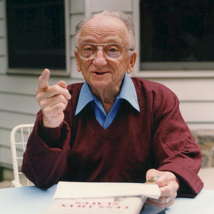 An old man sitting at a table with his hand raised.